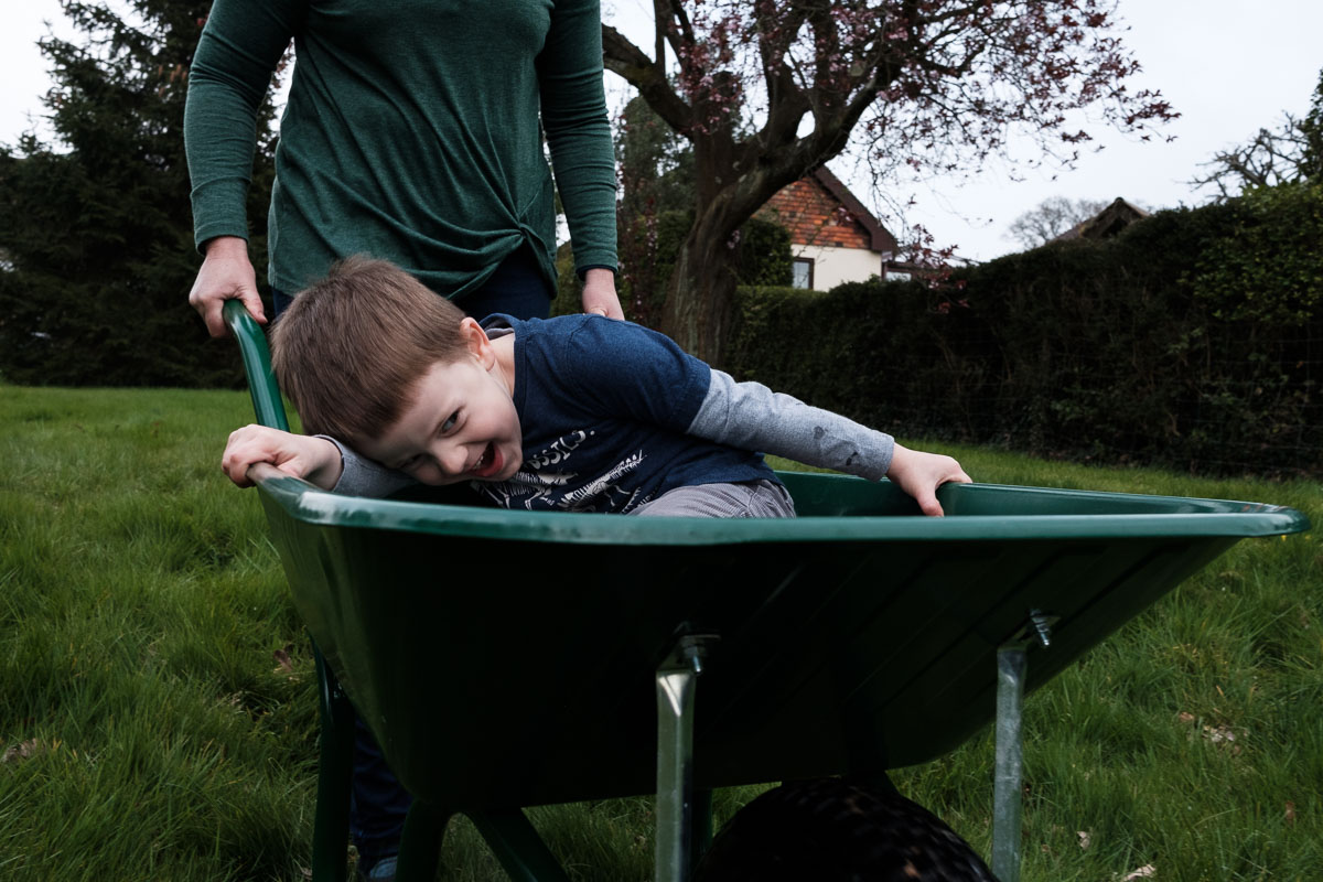 Child smiling whilst riding on a wheelbarrow pushed by mother.