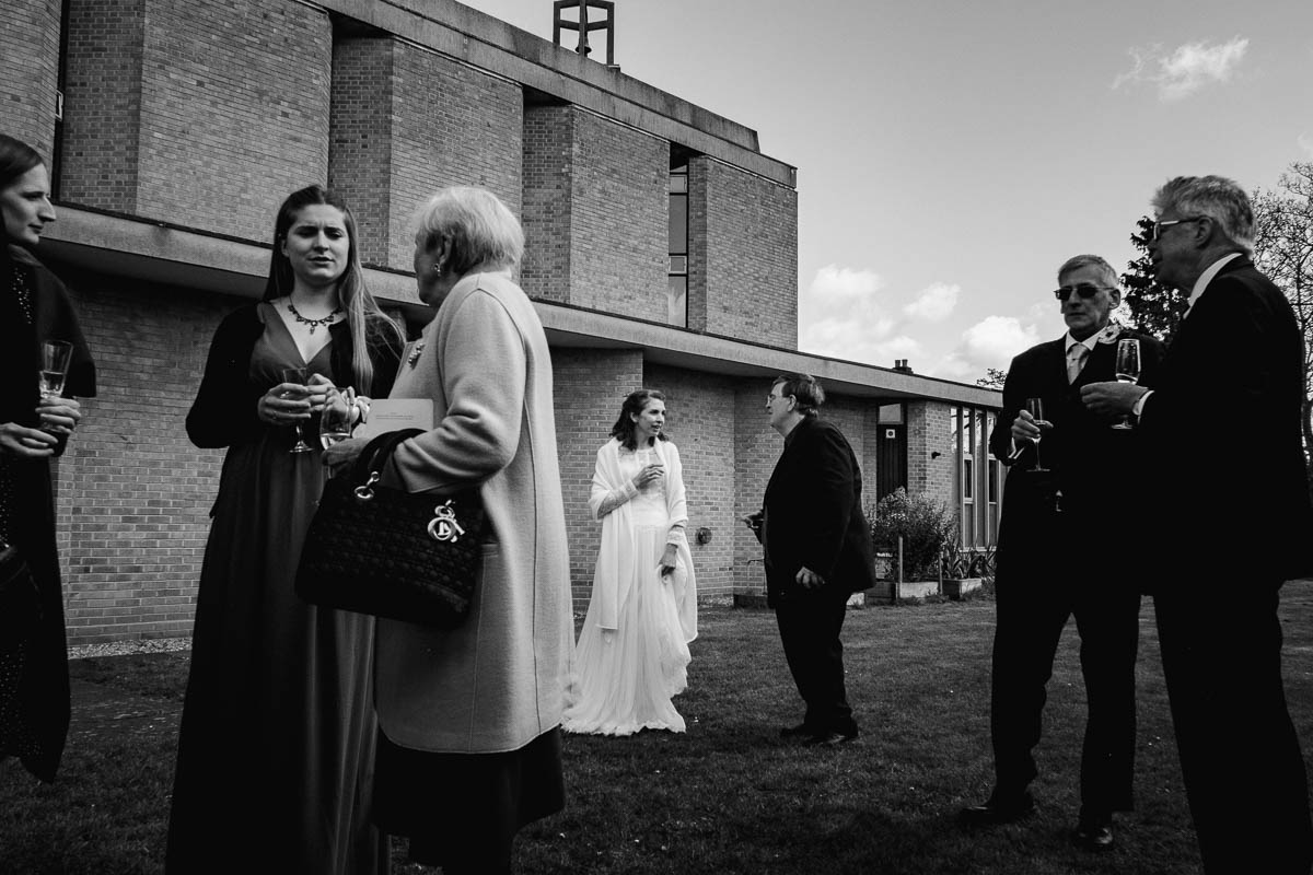 Bride stands in centre with guests outside church with drinks