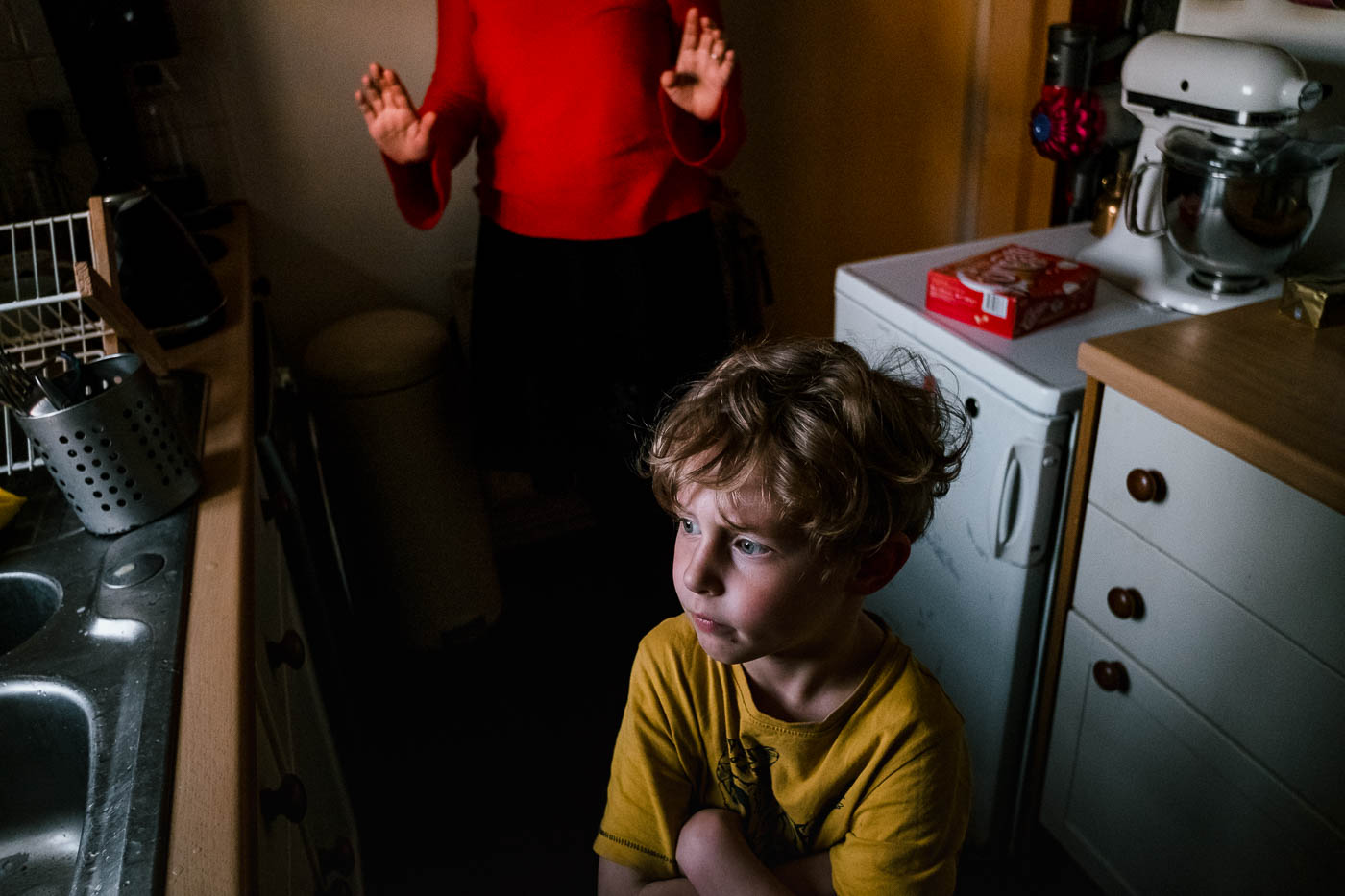 Boy crossing arms in kitchen with mother behind